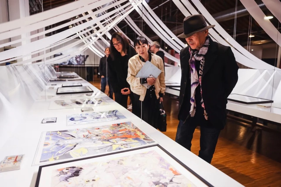 Amano with visitors at Milan &mdash; the artist viewing framed artworks alongside attendees during the exhibition opening