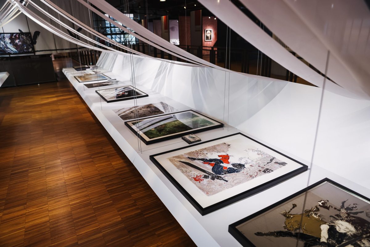 Framed original artworks suspended above display tables &mdash; the exhibition's POLI.design installation places works in dialogue across the gallery space