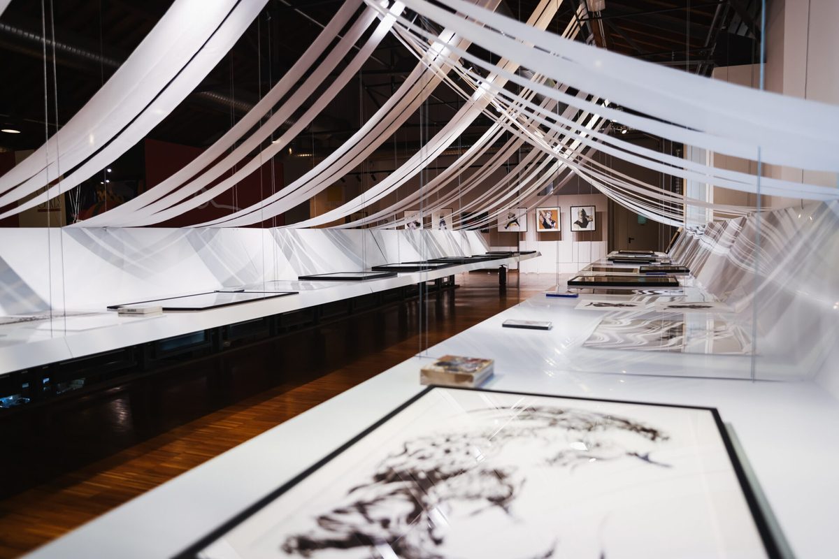 Milan venue installation &mdash; framed works displayed under POLI.design's dramatic white ribbon canopy at Fabbrica del Vapore