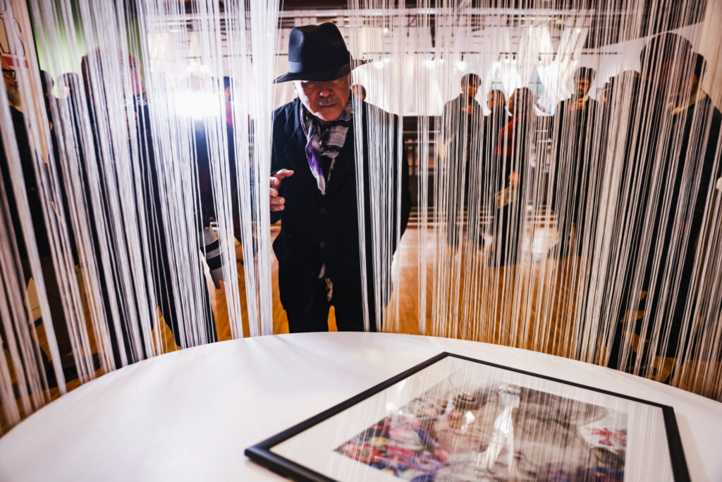 Amano viewing artwork through the exhibition's white string curtain installation &mdash; a design element by POLI.design connecting the gallery sections
