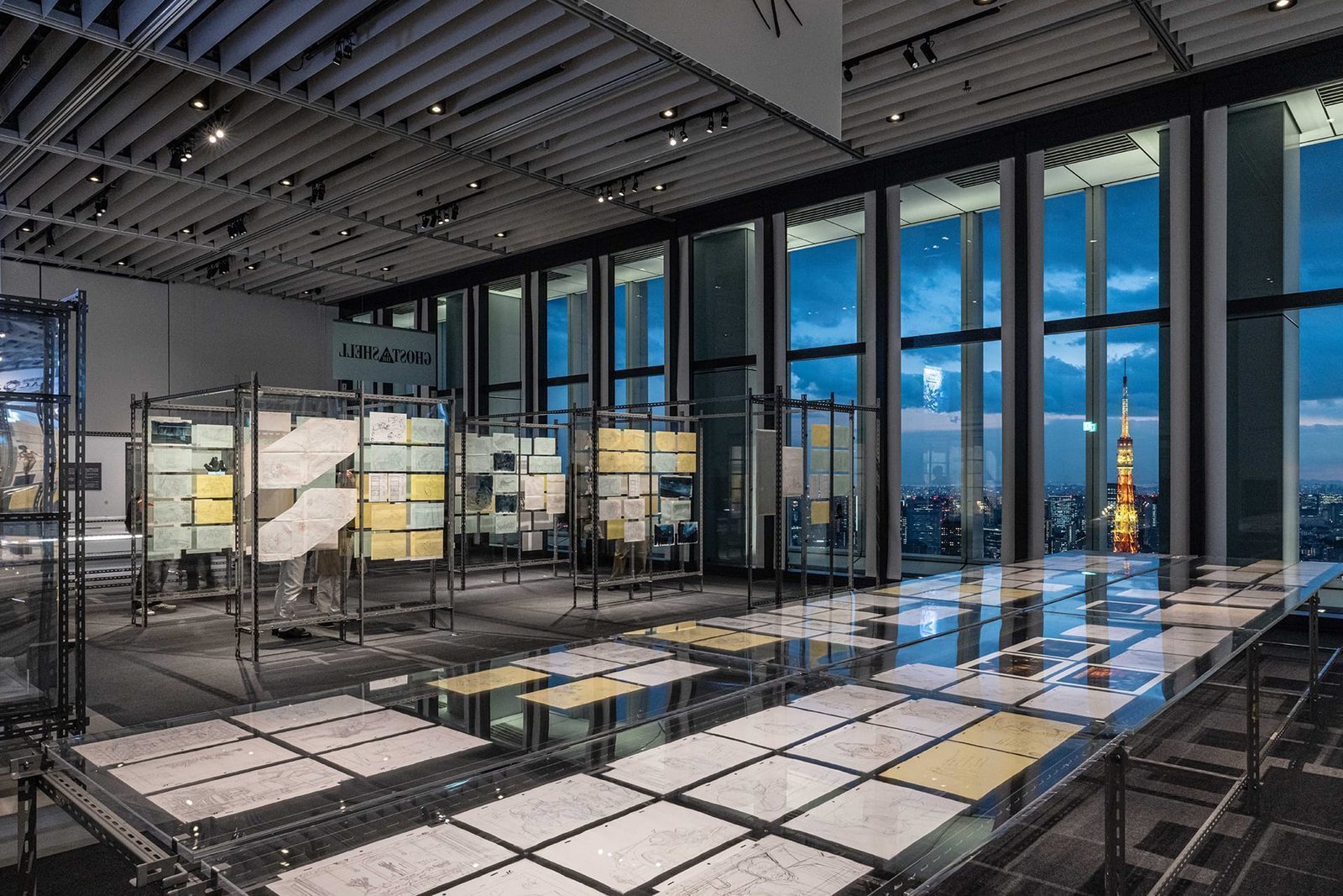 Gallery B (Dig) — 1,600+ production materials displayed on metal racks with Tokyo Tower visible through floor-to-ceiling windows. Original drawings, storyboards, layouts, cels spanning Oshii, Kamiyama, and the 2026 Science SARU series.
