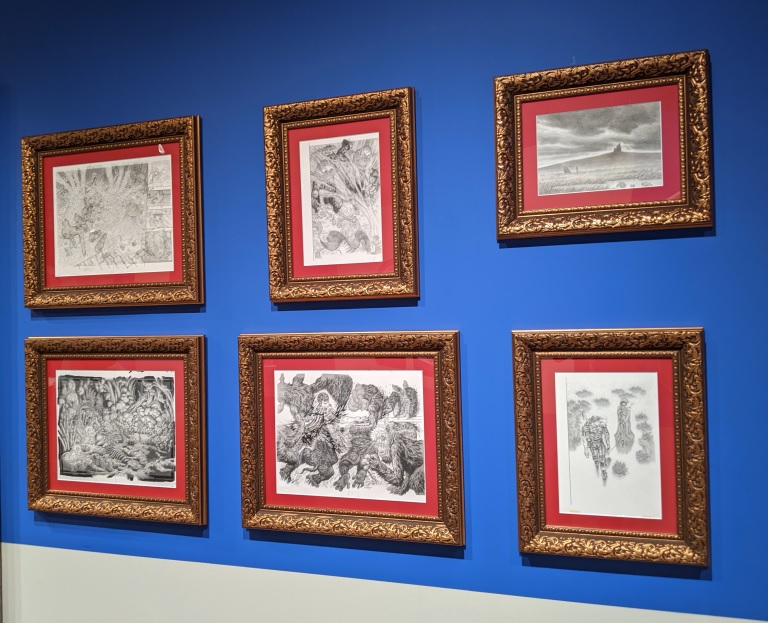 Original manuscripts gallery &mdash; six framed pages in gold frames on blue wall, showing Miura's obsessive hatching at exhibition scale