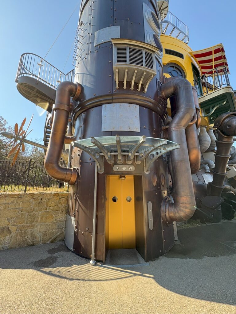 Castle entrance &mdash; the copper-clad cylindrical structure with pipes, riveted panels, balconies above, and the yellow door at ground level