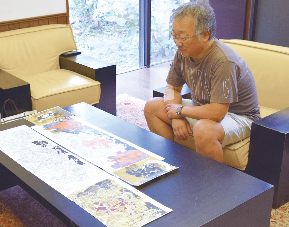 Otomo reviewing the clay maquette with CREARE artisans &mdash; reference photos and the <em>Kaba 2</em> illustration spread across the table