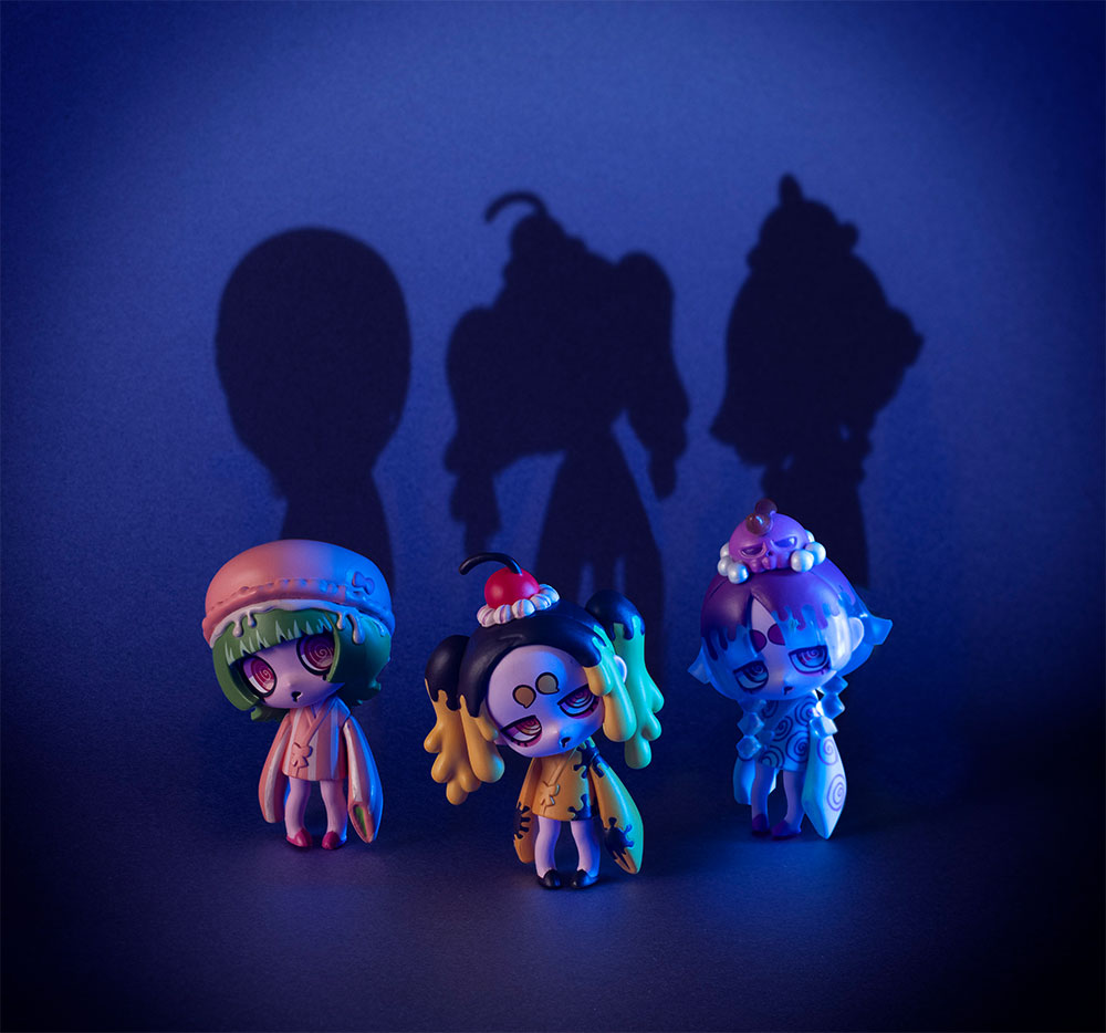 Three figures cast long shadows on dark blue. Abotoma's design reads differently under dramatic lighting — the sweetness recedes, the wrongness stays.